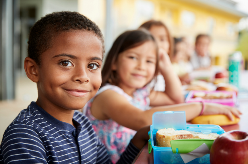Group of children sitting down at long table outside to eat lunch at school.
